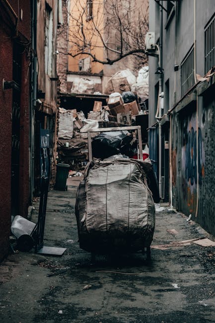 A disorganized collection of overflowing rubbish bins and scattered waste on a paved sidewalk in an urban environment, with various types of discarded materials including cardboard boxes, plastic bags