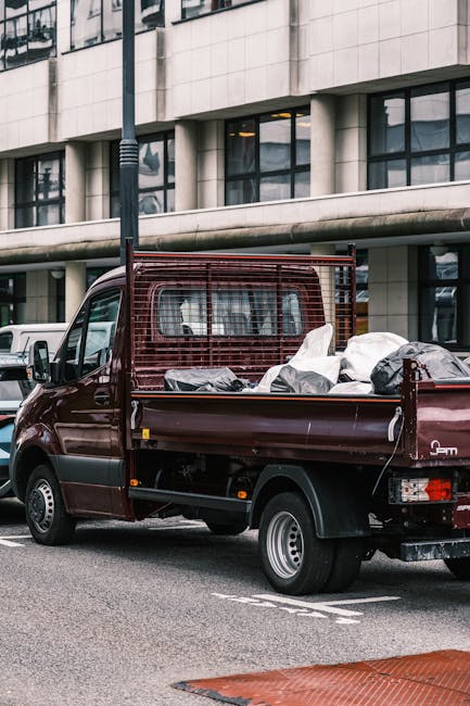 A dark red flatbed pickup truck is parked on an urban street in front of a multi-storey office building with large glass windows and a stone facade. The truck's cargo area is loaded with various types