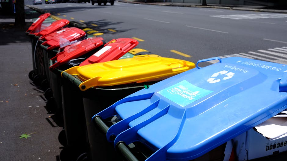 A row of multiple wheeled rubbish bins lined up along the edge of an asphalt street pavement, each bin featuring a different colour lid—red, yellow, and blue—and made of durable plastic with smoot